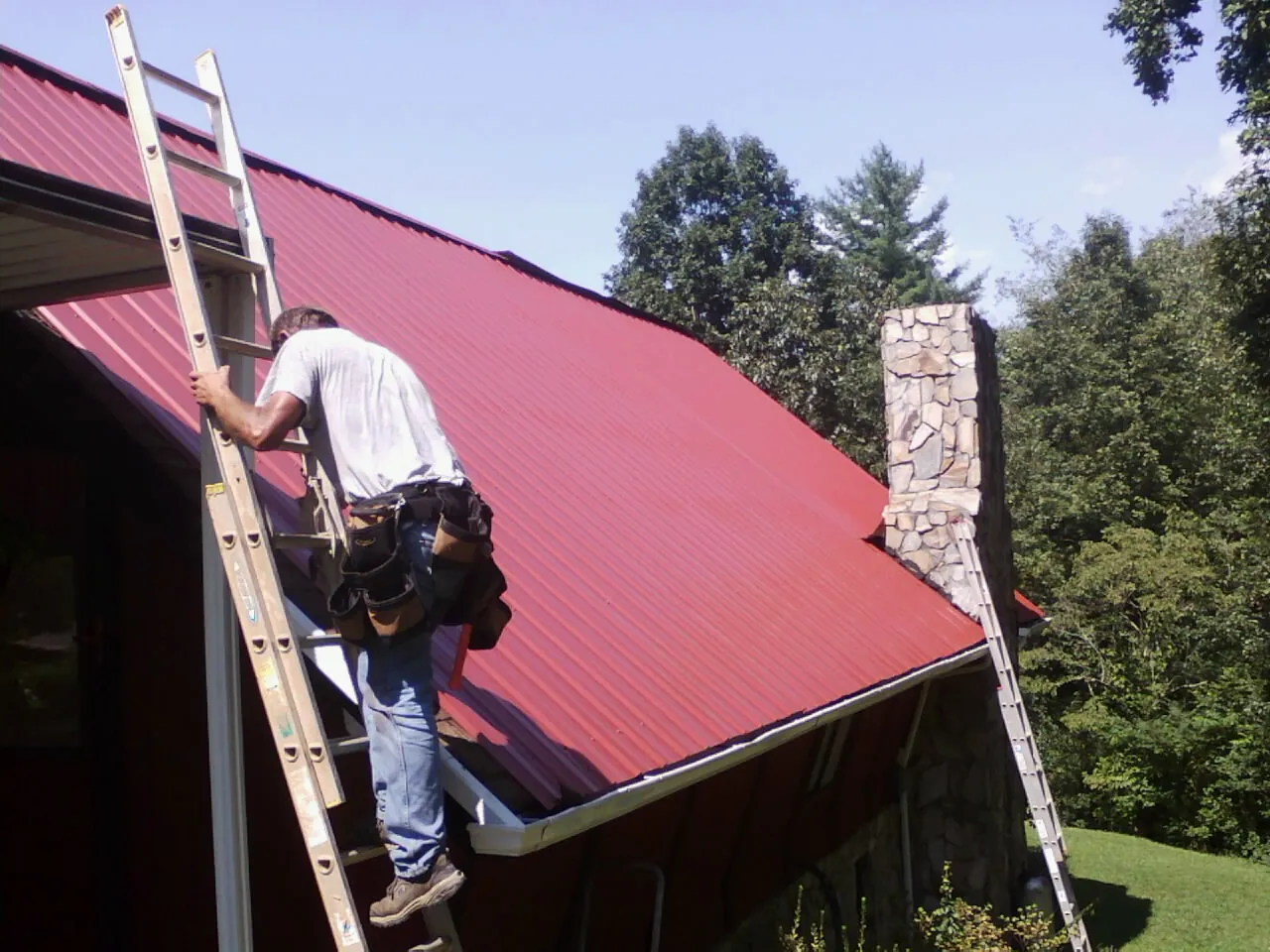 Roofer on a ladder beside a red metal roof and stone chimney — Soffit & Fascia Repair by Roofing Brothers in Brookdale